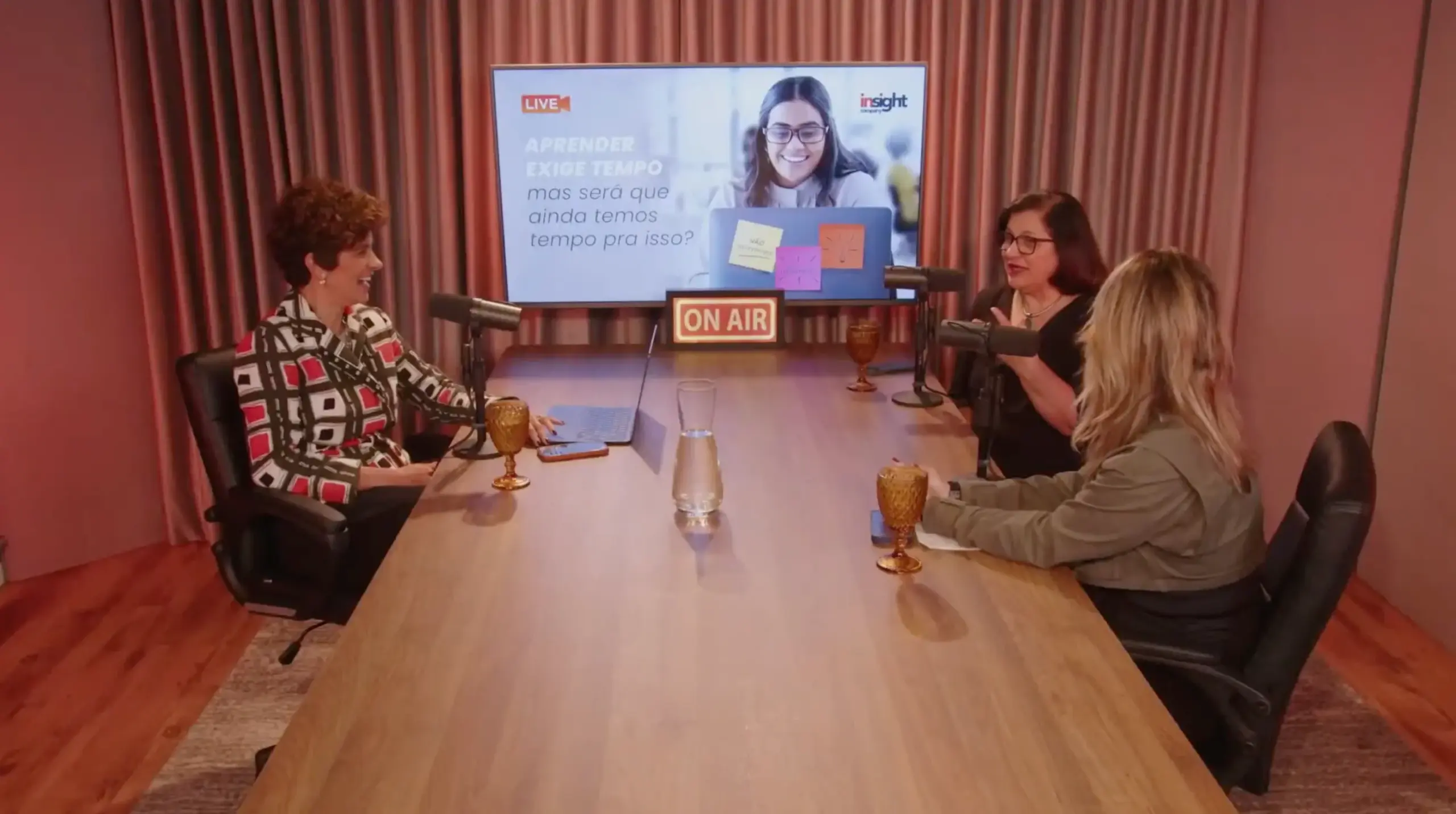 Três mulheres participam de conversa em estúdio, com microfones e tela ao fundo sobre aprendizagem e tempo.